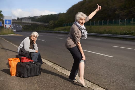Women, road and funny hitch hiker for travel in retirement, lift and senior friends in city with bags for adventure. Elderly, thumbs up and tired by roadside with comic and stop transport to commute.