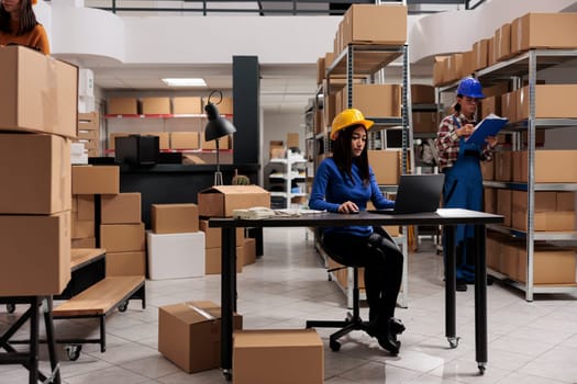 Shipment manager in helmet supervising warehouse operations on laptop