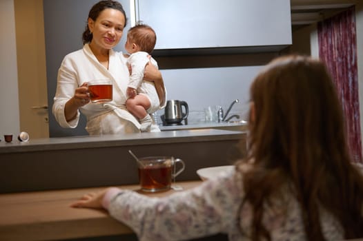 Authentic portrait of a young woman carrying her kid and having coffee in the morning at home with her daughter
