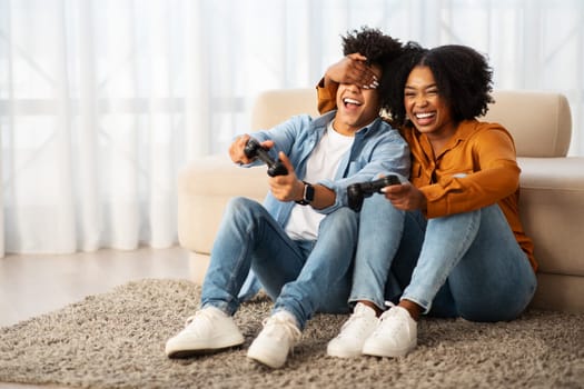 A woman playfully covers a man's eyes while they both laugh and hold game controllers