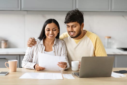 Happy hindu couple holding papers, calculating domestic bills, kitchen interior