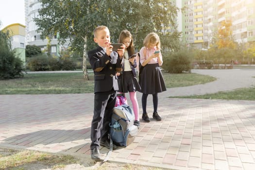 Children of elementary age with smartphones, backpacks, outdoor background.