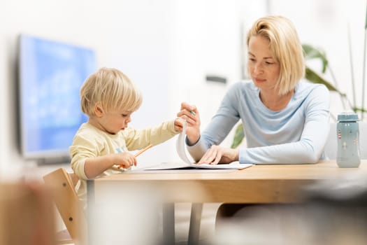 Caring young Caucasian mother and small son drawing painting in notebook at home together. Loving mom or nanny having fun learning and playing with her little 1,5 year old infant baby boy child.