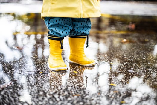Small infant boy wearing yellow rubber boots and yellow waterproof raincoat standing in puddle on a overcast rainy day. Child in the rain.