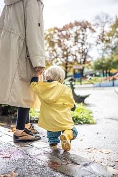 Small bond infant boy wearing yellow rubber boots and yellow waterproof raincoat walking in puddles on a overcast rainy day holding her mother's hand. Mom with small child in rain outdoors.