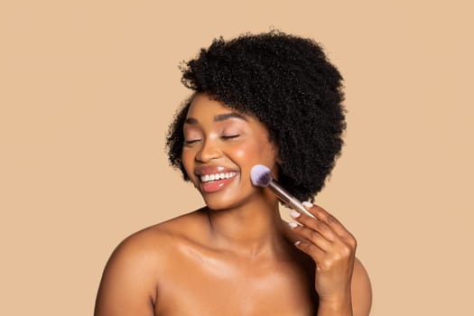 Happy black woman applying blush, closed eyes, beige backdrop