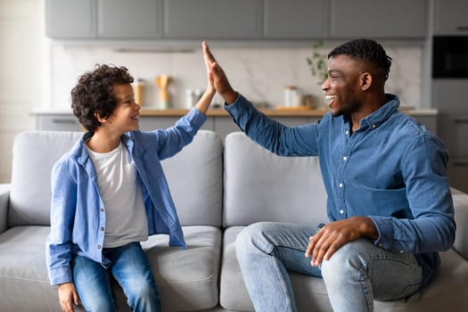 Smiling black boy gives his father high-five while sitting together on couch, both looking at each other with expressions of happiness and achievement