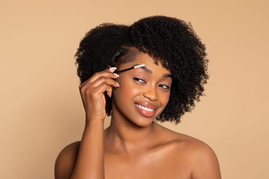 Young black woman grooming eyebrows, smiling, neutral backdrop