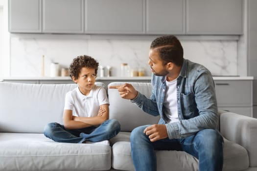 Angry african american father scolding upset little son at home, preteen black boy sitting on couch with arms crossed, dad and male kid having quarrel, suffering misunderstanding, closeup