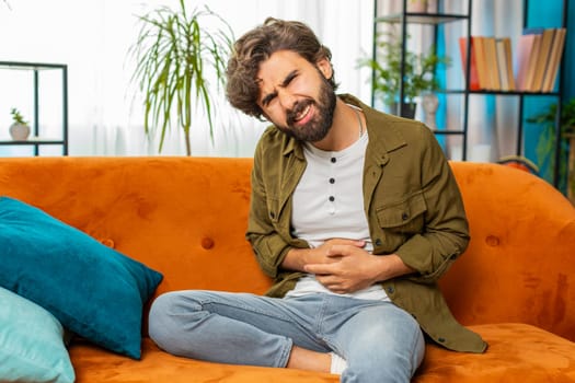Arabian man sits on sofa feeling sudden strong abdominal stomach ache, gastritis problem, diarrhea