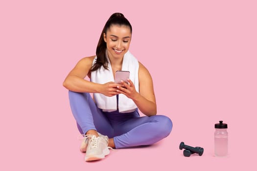 Content woman in purple workout outfit sits with smartphone, water bottle and dumbbells, taking break from exercise on pink background