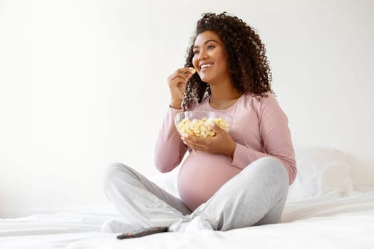 Happy black expectant mother in pink shirt enjoying bowl of popcorn