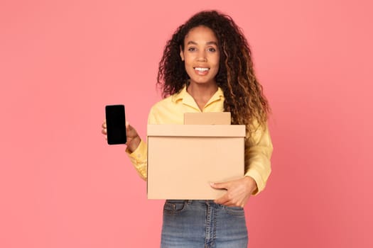 Young black woman with box parcels and blank phone screen on pink background