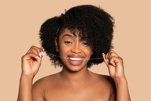 Joyful black woman flossing teeth, curly hair, beige background