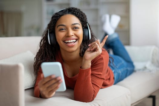 Cheerful Young Black Woman Wearing Wireless Headphones Relaxing With Smartphone At Home