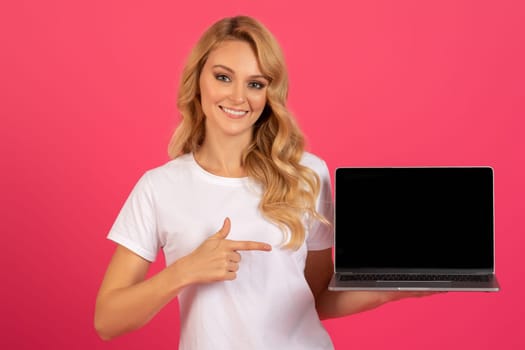 Happy blonde woman pointing at laptop with empty screen, studio