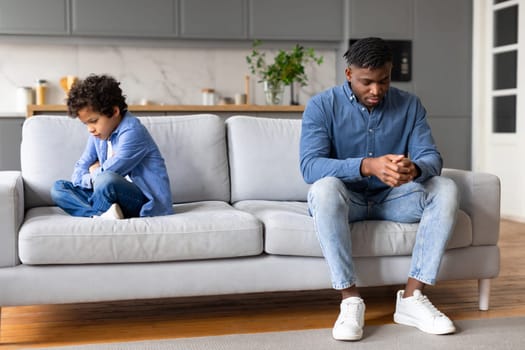 Upset black father and son sitting apart on couch, having disagreement