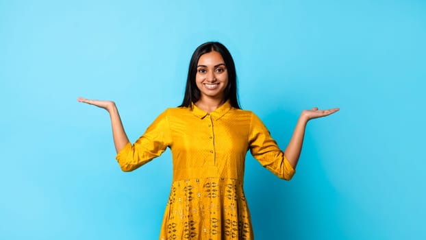 Smiling Hindu woman making scales gesture with hands, blue background