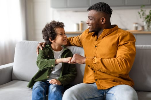 Black father and son share fist bump and smile on sofa
