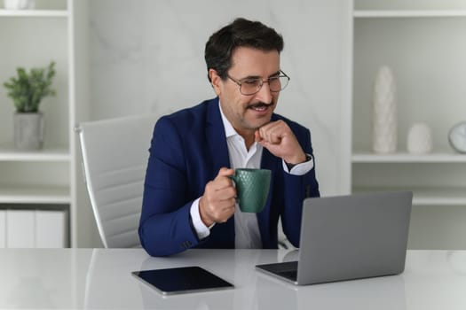 Businessman in blue suit enjoys a cup of coffee while reading on his laptop