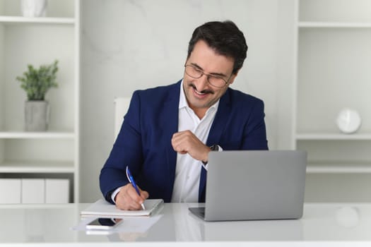 Cheerful businessman in a blue suit punches the air with joy while working on his laptop at a white desk