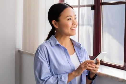 Dreamy millennial chinese woman with smartphone next to window