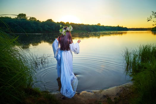 Adult mature brunette woman in a white dress, sundress and a wreath of flowers in summer in water of river or lake in evening at sunset. Celebration of the Slavic pagan holiday of Ivan Kupala