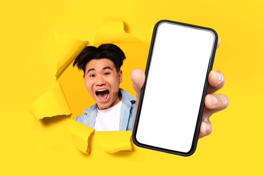 Excited asian guy holding mobile phone breaking through yellow paper