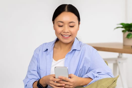 Positive korean lady enjoying weekend at home, using smartphone