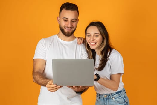 A man and woman in white shirts and blue jeans happily share a view on a laptop