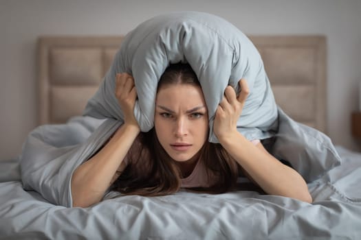 Irritated woman covering ears with pillow in bed