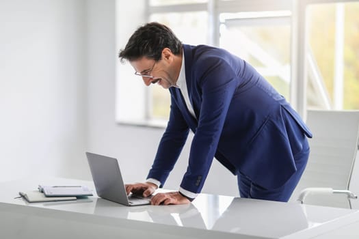A caucasian cheerful busy mature businessman in a blue suit leans over his laptop, laughing heartily while working in a modern, brightly lit office. Work, business with gadget