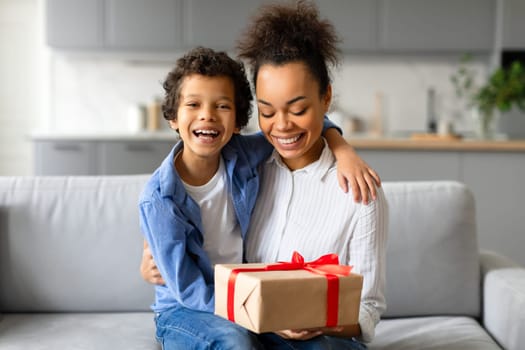 Joyful black mother and son with gift box on sofa