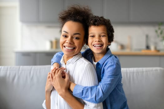Joyful black mother and her preteen son with curly hair embrace warmly, sharing bright smile in loving moment at home sitting together on the couch
