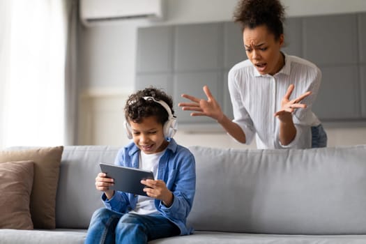 Angry black mother gestures with concern towards her son, who is engrossed in tablet with headphones on, oblivious to her frustration, home interior