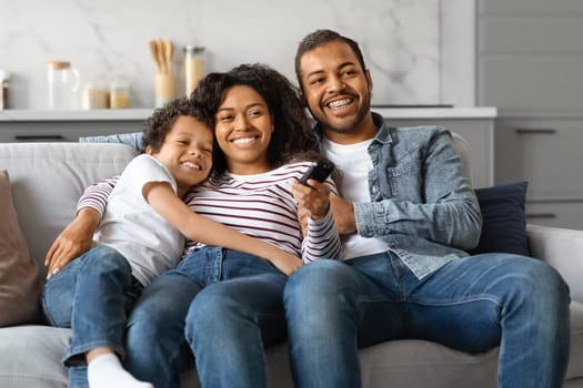 Joyful black family of three relaxing on sofa with remote controller