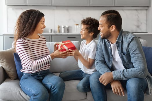 Holiday Present. Excited Black Woman Receiving Wrapped Gift Box From Her Husband And Little Son At Modern Home Interior, African American Family Celebrating Birthday And Having Fun Together