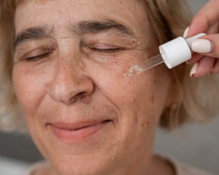 Close-up portrait of an old woman applying hyaluronic acid serum with a pipette. Anti-aging face care.
