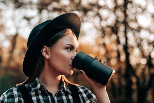 Hipster woman drinking hot coffee or tea, thermos, golden park, forest in autumn
