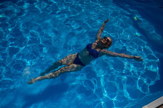 An elderly woman in sunglasses swims on her back in the pool. Vacation in retirement.