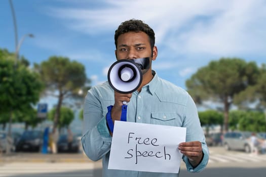 Young man with taped mouth trying to speak into megaphone standing outdoors.