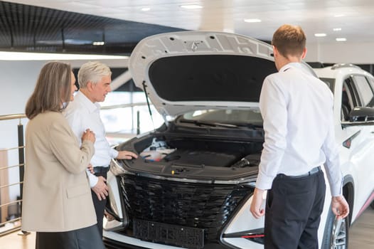 A salesman demonstrates a car with an open hood to an elderly Caucasian couple in a car dealership.