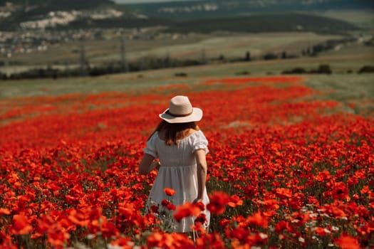 Field poppies woman. Happy woman in a white dress and hat stand with her back a blooming field of poppy. Field of blooming poppies.