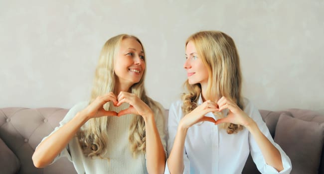 Happy smiling caucasian middle aged mother with adult daughter together showing heart shaped gesture sign with hands and looking at each other at home