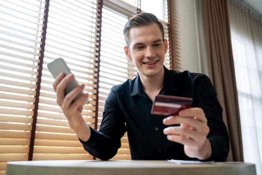 Young man sitting at table using online payment app and digital wallet on smartphone to pay with credit card. E commerce shopping and modern purchasing via mobile internet. Unveiling