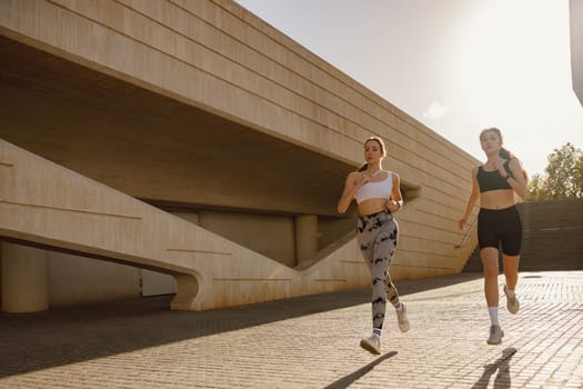Two active women athlete running on sunny morning side by side on modern buildings background