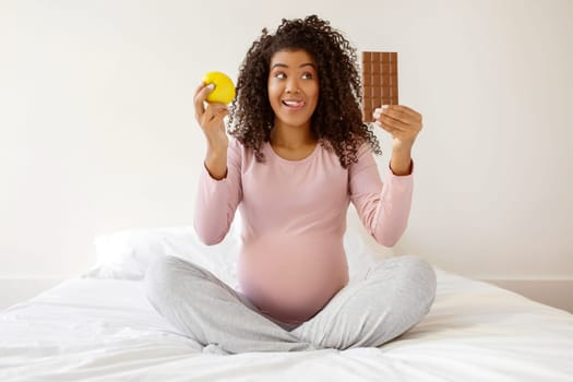 Cheerful black expectant mother sitting on bed, comparing apple to chocolate bar