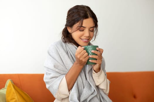 Smiling young woman wrapped in blanket enjoying cup of tea at home