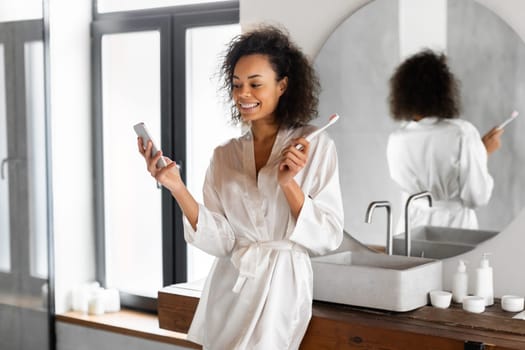 Happy African American woman checks phone brushes teeth in bathroom
