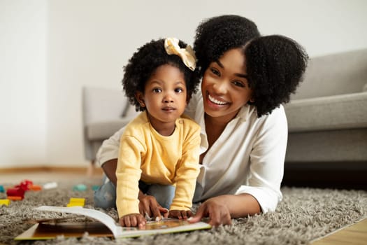 Happy african american young mother and little daughter playing at home, laying on floor, reading books, mom entertaining her toddler kid with various toys. Child development